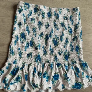 Blommig kjol med volang - Superfin blommig kjol i vitt med blå blommor och volangdetalj längst ner. Perfekt för sommardagar eller en mysig vårutflykt. Kjolen har en smockad design som ger en bekväm passform. Passar perfekt till en enkel topp och sandaler för en avslappnad look.