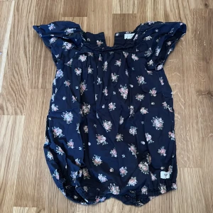 Blommig byxdress från Newbie - Söt blommig byxdress från Newbie i mörkblått med små rosa och vita blommor. Byxdressen har korta ärmar och knappar i ryggen för enkel påklädning. Perfekt för de små under vår och sommar.