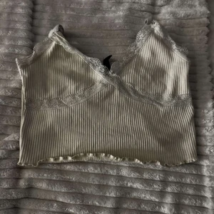 Beige ribbad linne med spetsdetaljer - Säljer en söt beige ribbad linne med spetsdetaljer vid halsringningen och axelbanden. Toppen är croppad och har en omlott-design framtill. Perfekt för varma dagar eller som ett lager under en tröja. ⚠️ ni betalar för frakt