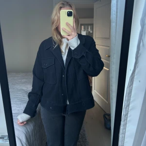 Svart jacka med fickor - Snygg svart jacka med knappar och stora fickor framtill. Perfekt för en stilren look. Jackan har långa ärmar och en klassisk krage.