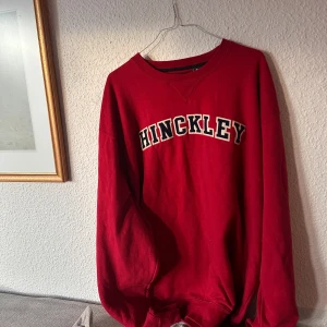 Röd sweatshirt med text - Snygg röd sweatshirt med texten 'HINCKLEY' i svart och vitt på bröstet. Tröjan har en klassisk rund halsringning och ribbade muddar vid ärmslut och nederkant. Perfekt för en avslappnad stil.