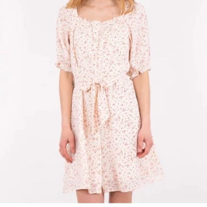 Blommig klänning från Neo Noir - Charmig blommig klänning från Neo Noir i en ljus nyans med små rosa blommor. Klänningen har korta ärmar och ett knytband i midjan för en smickrande passform. Perfekt för somriga dagar!