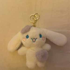 cinnamoroll sanrio nyckelring - En söt, vit och fluffig nyckelring i form av en kanin med långa öron. Den har blå broderade ögon och en glad mun, samt små rosa hjärtan på kinderna. Nyckelringen har en guldfärgad kedja och ring.