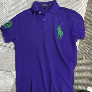 Lila pikétröja från Ralph Lauren - Säljer en snygg lila pikétröja från Ralph Lauren i slim fit. Den har korta ärmar och en grön broderad logga på bröstet samt en grön siffra på ärmen. Perfekt för en stilren och sportig look!