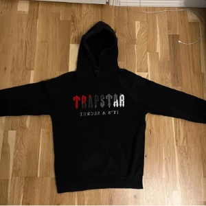 Svart hoodie från Trapstar - Säljer en svart hoodie från Trapstar med texten 'It's a Secret' i vitt och rött på framsidan. Den har en klassisk design med en stor ficka framtill och en bekväm huva. Perfekt för en avslappnad stil. Pris går att diskuteras:)