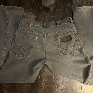 Grå jeans från Wrangler - Säljer ett par grå jeans från Wrangler i bra skick. De har en klassisk femficksdesign med snygga sömmar på bakfickorna och en läderpatch med logga. Perfekta för vardagsbruk och passar till det mesta. Dragkedja framtill och normal passform.