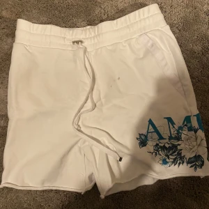 Vita shorts från Amiri - Snygga vita shorts från Amiri med blått blommönster och textdetalj. De har snörning i midjan och en bakficka. Perfekta för en avslappnad stil. Använd ett fåtal gånger men tyvärr har den några svarta fläckar som jag själv inte kunnat få bort 
