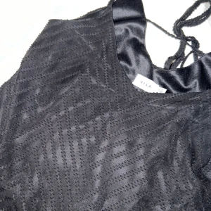 Svart spetslinne från Vila - Snyggt svart spetslinne från Vila med tunna axelband och ett diskret mönster. Perfekt för en stilren look. Linnet har en elegant knytning i ryggen.