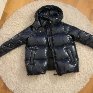 Svart glansig dunjacka med huva - Snygg och varm svart dunjacka från Tommy Hilfiger med glansig finish och huva. Jackan har dragkedja och praktiska fickor. Perfekt för kyliga dagar. Den har små märken vid ärmarna men inget som syns tydligt, kom privat för bilder på defekterna🥰 Säljer pågrund av att den är för stor💗