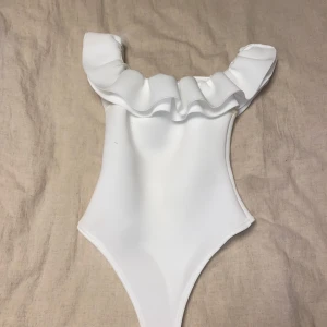 Vit body med volang från Boohoo - Säljer en superfin vit body från Boohoo med volangdetaljer runt axlarna. Perfekt festtopp! Den är i nytt skick och har en elegant och stilren design. Passar perfekt för dig som vill ha något unikt och trendigt!