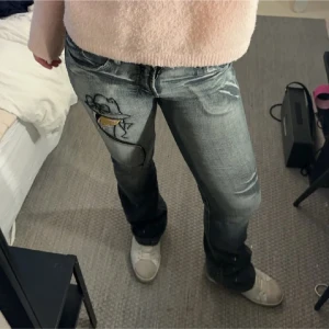 Grå jeans med broderad detalj - Säljer ett par grå jeans med en cool broderad detalj på ena benet. De har en klassisk femficksdesign och en bekväm passform. Perfekta för en avslappnad vardagslook!