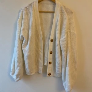 Vit stickad kofta med knappar - Mysig vit stickad kofta med stora träknappar framtill. Perfekt för kyliga dagar, den har en avslappnad passform och långa ärmar. Koftan är i bra skick och passar både till vardags och lite finare tillfällen. Ett måste i garderoben för höst och vinter!