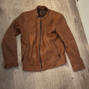 Brun skinnjacka från Jack & Jones - Säljer en snygg brun skinnjacka från Jack & Jones i storlek L. Men liten i storlek så mer som medium. Jackan har en dragkedja framtill och två fickor med dragkedja. Perfekt för höst och vår, med en stilren design som passar till det mesta. Använd men i mycket bra skick.
