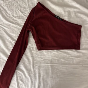 oneshoulder topp - Säljer en snygg vinröd oneshoulder topp från Boohoo i ribbat material. Toppen är croppad och har en lång ärm, perfekt för en utekväll eller fest. Den är i mycket bra skick och passar perfekt till höga jeans eller kjol. Supertrendig och bekväm! Storlek XS, pris 30:-
