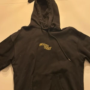 Svart hoodie från Vailent - Säljer en svart hoodie från Vailent med texten 'GOOD DAY' i gult på bröstet och en stor grafisk design på ryggen med samma text, en glad blomma och en hand. Perfekt för en avslappnad stil.