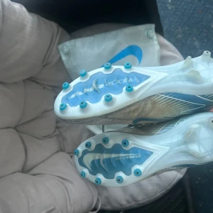 Nike fotbollsskor med blå detaljer använda i tre månader  - Snygga fotbollsskor från Nike med blå detaljer och snörning. Skorna har en modern design med en kombination av syntetmaterial och en slitstark sula för optimalt grepp på planen.