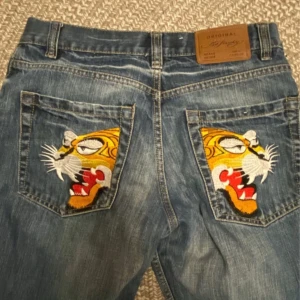 Blå jeans med tigerbroderi - Snygga blå jeans med unikt tigerbroderi på bakfickorna. De har en klassisk femficksdesign och en rak passform. Perfekta för dig som vill sticka ut med en cool detalj. Vet ej om de är äkta. Midjan rakt över: 36 cm. Innerben: 76cm. Benvidd längst ner: 20cm.