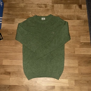 Grön stickad tröja från Lacoste - Säljer en snygg grön stickad tröja från Lacoste med det klassiska krokodilmärket på bröstet. Tröjan har långa ärmar och en rund halsringning. Perfekt för kyligare dagar!