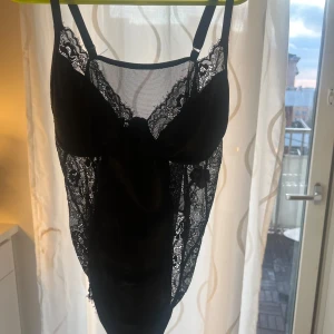Svart spetsbody - Elegant svart body med spetsdetaljer och justerbara axelband. Den har en vacker kombination av satin och spets som ger en lyxig känsla. Perfekt för speciella tillfällen.