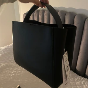 Svart väska - Stilren svart väska i syntetmaterial. Väskan har en enkel och elegant design med en justerbar axelrem. Aldrig använd , prislapp kvar 