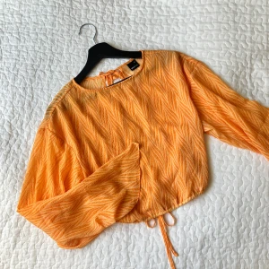 Orange blus med knytband - Säljer en snygg orange blus med ett subtilt mönster och knytband i ryggen. Blusen har långa ärmar och en luftig passform, perfekt för varmare dagar.