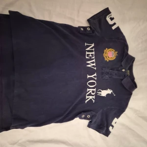 Mörkblå pikétröja från Polo Ralph Lauren - Snygg mörkblå pikétröja från Polo Ralph Lauren med texten 'New York' och ett broderat emblem på bröstet. Tröjan har en klassisk krage med knappar och stora siffror på ärmarna. Perfekt för en sportig look!