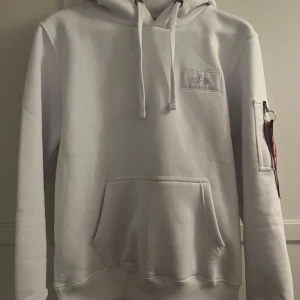 Vit hoodie från Alpha Industries - Säljer en stilren vit hoodie från Alpha Industries med stor svart logga på ryggen. Hoodien har en klassisk känguruficka och en cool detalj med 'Remove Before Flight' band på ärmen. Perfekt för en casual look. Varan är ny med prislapp kvar, så aldrig använd. Köparen betalar frakt och den skickas inom 1-2 dagar. ✅