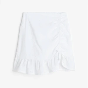 Vit kjol med volang och knyt - Söt vit kjol med volangdetalj och knyt på sidan. Kjolen har ett broderat mönster och är perfekt för sommarens varma dagar. Den har en kort längd och en omlott-design som ger en fin passform. HELT SLUTSÅLD ÖVERALLT 💕