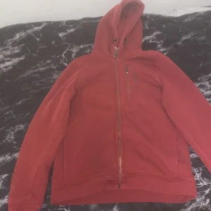 Röd fleecejacka från Sail Racing - Säljer en röd fleecejacka från Sail Racing med dragkedja och huva. Jackan har en praktisk bröstficka med dragkedja och broderad logotyp. Perfekt för kyliga dagar.