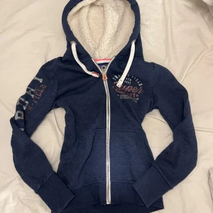 Mörkblå hoodie från Superdry - Säljer en mörkblå hoodie från Superdry med dragkedja och fodrad huva. Tröjan har tryck på ärmen och bröstet samt praktiska fickor framtill. Perfekt för kyliga dagar!