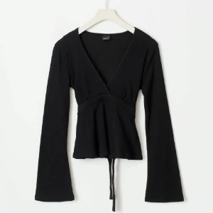 Svart ribbad omlottopp med knytning i ryggen - Snygg svart ribbad omlottopp med v-ringning och långa ärmar. Toppen har knytband i midjan för en justerbar passform. Perfekt för en stilren look. I mycket bra skick💗