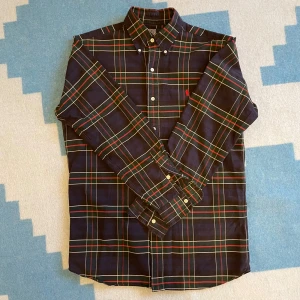 Rutig skjorta från Polo Ralph Lauren - Snygg rutig skjorta från Polo Ralph Lauren i mörkblått med gröna och röda ränder. Tillverkad i 100% bomull, perfekt för en stilren look. Skjortan har långa ärmar och knappar framtill.