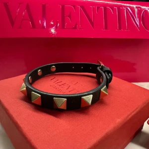 Svart armband med nitar från Valentino - Snyggt svart armband från Valentino dekorerat med guldiga pyramidformade nitar. Perfekt accessoar för att ge en edgy touch till din stil. Kommer i originalförpackning med tillhörande påse och kort.