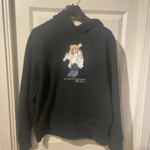Svart hoodie från Ralph Lauren med Polo Bear Bear - Säljer en svart hoodie från Ralph Lauren med det ikoniska Polo Bear-motivet på framsidan. Tröjan har en rymlig ficka framtill och en bekväm huva. Perfekt för en avslappnad stil.