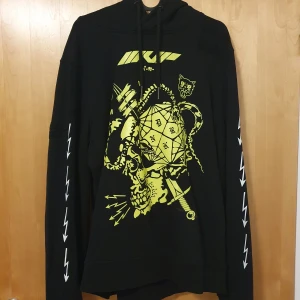 Svart hoodie med grafiskt tryck från DRKN - Säljer en svart hoodie från DRKN med gult grafiskt tryck fram och bak. Tröjan har långa ärmar med vita blixtar längs sidorna och en bekväm huva med dragsko. Perfekt för dig som gillar unika och iögonfallande plagg!