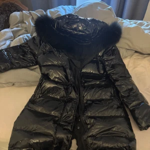 Svart dunjacka med pälskrage - Snygg svart dunjacka med glansig finish och en mysig pälskrage. Jackan har en lång modell med dragkedja och knappar för extra värme. Perfekt för kalla vinterdagar. Storlek M men liten i storleken skulle nog säga S 