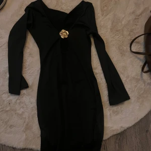 Svart långärmad klänning med gulddetalj - Elegant svart klänning med långa ärmar och en vacker gulddetalj framtill. Perfekt för en stilren look.