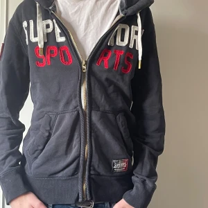 Superdry zip hoodie - Säljer en svart hoodie från Superdry med texten 'SUPERDRY SPORTS' i vitt och rött på framsidan. Hoodien har en dragkedja och två fickor framtill samt en broderad logga på ärmen. Perfekt för en avslappnad stil! NYPRIS 1500 vårt 400