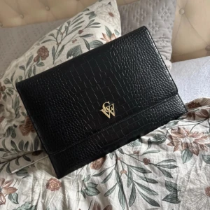 Svart kuvertväska med krokodilmönster - Elegant svart kuvertväska med krokodilmönster och guldfärgad logotyp på framsidan. Väskan har ett stilrent innerfoder med monogrammönster och stängs med magnetknappar.