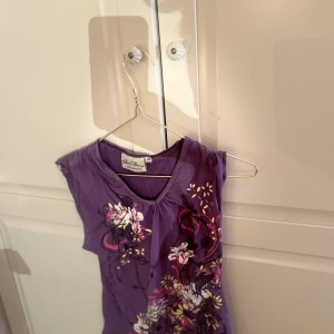 Lila blommig topp  - Säljer en lila topp med blommigt mönster i vitt och rosa. Toppen har en v-ringad hals och är ärmlös, perfekt för varma dagar. Passar bra till både jeans och kjol. 🌸