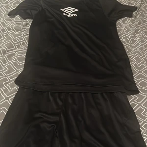 Svart fotbollströja från Umbro med umbro shorts - Säljer en stilren svart fotbollströja från Umbro. Tröjan är kortärmad och perfekt för träning eller match. Ett par byxor ingår också.