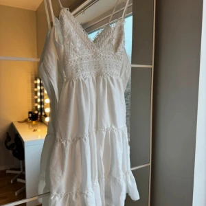 Vit klänning med spetsdetaljer - Charmig vit klänning med smala axelband och v-ringad spetsdetalj. Klänningen har en luftig passform med volanger och är perfekt för sommarens varma dagar.