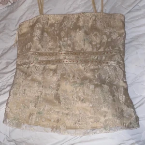 Beige blommig tubtopp från Vila - Säljer en elegant beige tubtopp från Vila med blommigt mönster och smala axelband. Toppen har fina detaljer med broderade blommor och en subtil glans. Perfekt för en stilren look.. använd endast en gång