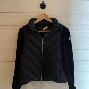 Svart dunjacka från Moncler - Snygg svart cardigan från Moncler med långa ärmar och dragkedja framtill. Jackan har ett quiltat mönster och moncler märket är broderat på ärmen. Perfekt för kyligare dagar. Använd fåtal gånger och är 8/10 i skick, det är bara att höra av er om något om jackan. Pris ej hugget i sten!