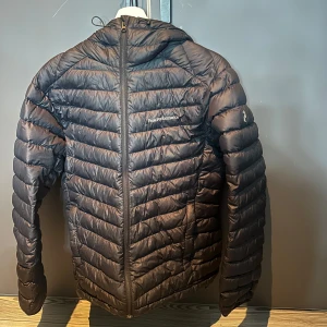 Svart dunjacka från Peak Performance - Snygg svart dunjacka från Peak Performance med dragkedja och huva. Perfekt för kyliga dagar med sin quiltade design som håller värmen. Jackan har en stilren look och är lätt att matcha med olika outfits.