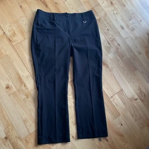 Svarta kostymbyxor från KappAhl - Snygga svarta kostymbyxor från KappAhl med hög midja och raka ben. Byxorna har en stilren design med knappar i midjan och hällor för bälte. Perfekta för en elegant look.