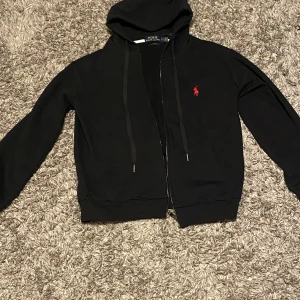 Svart hoodie från Polo Ralph Lauren - Säljer en stilren svart hoodie från Polo Ralph Lauren med en broderad röd logotyp på bröstet. Hoodien har dragkedja och långa ärmar, perfekt för en casual look. Passar bra till både jeans och joggers.