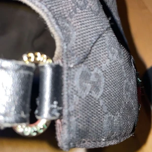 Svart keps från Gucci - Snygg svart keps från Gucci med diskret mönster och klassiska ränder i grönt och rött på sidan. Kepsen har en justerbar rem bak för perfekt passform. Tillverkad i en blandning av bomull och polyester. 