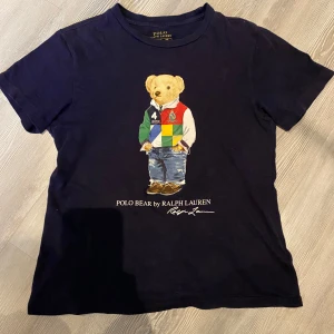 Svart t-shirt med Polo Bear från Ralph Lauren - Säljer en svart t-shirt från Ralph Lauren med en ikonisk Polo Bear-tryck på framsidan. Björnen har en färgglad outfit med jeans och sneakers. Perfekt för en avslappnad stil. 🐻