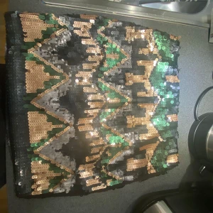 Paljettkjol med geometriskt mönster - Snygg kortkjol täckt av paljetter i geometriskt mönster med färgerna svart, grön, guld och silver. Perfekt för att sticka ut på festen! Kjolen har en tight passform och är ett riktigt statement-plagg.